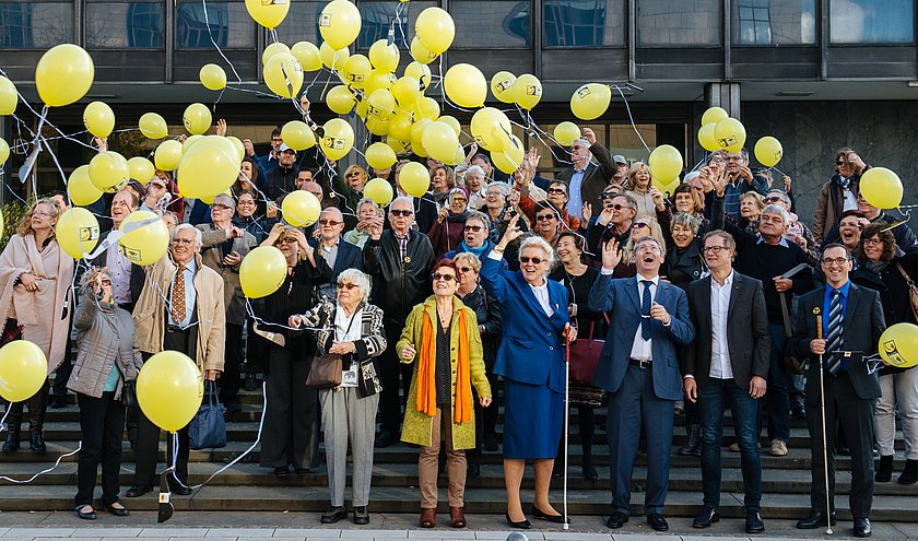 Viele Menschen stehen aufgereiht auf einer Treppe und lassen gelbe Luftballons steigen. 