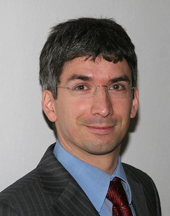 Portrait eines Mannes mit kurzen schwarzen Haaren, randloser Brille, blauem Hemd, roter Krawatte und grauem Jacket 