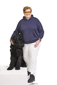 Heike Ferber trägt einen blauen Pullover und eine weiße Hose. Neben ihr sitzt ihr Blindenführhund.