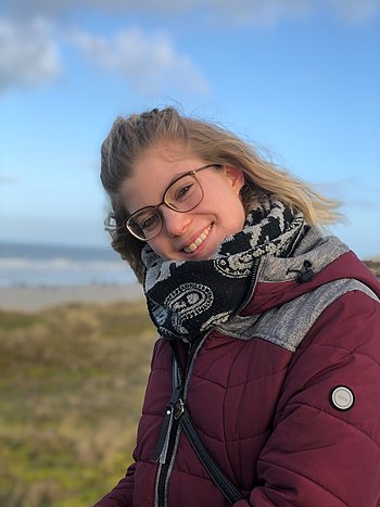 Portrait einer jungen Frau mit schulterlangen blonden Haaren, Brille und roter Winterjacke. Im Hintergrund Dünen und Meer.