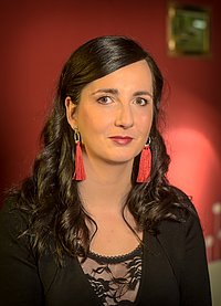 Jennifer Sonntags Portrait zeigt eine june Frau mit langen dunken Haaren, schwarzer Kleidung und roten Quastenohrringen vor einer roten Zimmerwand. Sie blickt direkt in die Kamera.