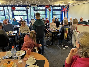 Eine Gruppe von Menschen sitzt in einem festlich dekorierten Raum bei Kaffee und Kuchen, während ein Junge eine Geschichte vorliest.