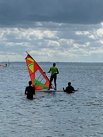 Zu sehen ist ein Teilnehmer beim Windsurfen. Er steht auf einem Surfbrett mit Segel auf dem Wasser. In unmittelbarer Nähe im Wasser befinden sich zwei Personen.