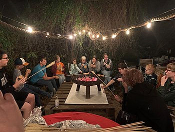 Für die Kinder und Jugendlichen gab es leckeres Stockbrot frisch über dem Lagerfeuer gebacken, am Strand des Ufercafes. Alle sitzen um eine große Feuerschale herum. Es ist dunkel, eine Lichterkette wirft gemütliches Licht auf die Gruppe.