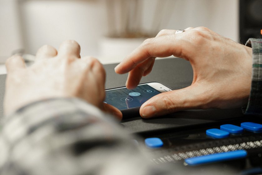 Großaufnahme von einer Hand, die über den Bildschirm eines Smartphones wischt. Davor liegt eine Braille-Zeile. 