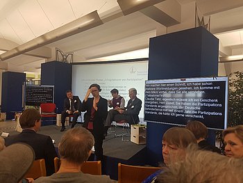 Auf einer Bühne sitzen die Teilnehmer einer Talkrunde. Eine Gebärdendolmetscherin übersetzt für die Zuschauer, von denen einige auf Stühlen sitzend von hinten zu sehen sind.