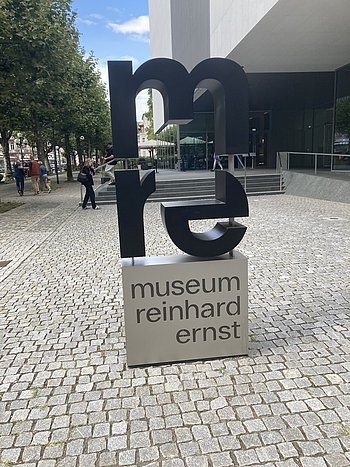 Eine Skulptur aus den schwarzen Anfangsbuchstaben des Museums mre auf einem silbernen Sockel mit dem Schriftzug Museum Reinhard Ernst 