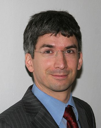 Portrait eines Mannes mit kurzen schwarzen Haaren, randloser Brille, blauem Hemd, roter Krawatte und grauem Jacket