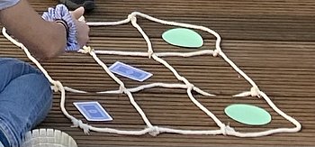 Zu sehen ist ein Spielfeld von Tic Tac Toe- auch Drei gewinnt genannt-, das aus Seil selber gebastelt wurde. Als Spielsteine werden eckige Spielkarten und rundes farbiges Papier verwendet.