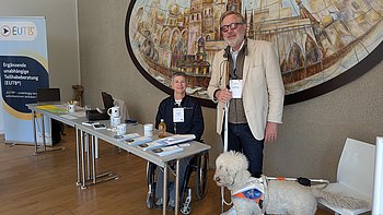 Claudia Posch von der EUTB Fachstelle Teilhabeberatung und Jörg-Michael Sachse-Schüler, Vorstandsmitglied von PRO RETINA Deutschland e.V. stehen hinter den Tischen am EUTB Stand. Hinter Ihnen ist ein klassisches Wandgemälde zu sehen. Das EUTB Banner ist auf der linken Seite. Jörg-Michael Sachse-Schüler (rechts von Claudia Posch, die auf einem Stuhl sitzt) hat seinen Assistenzhund dabei (rechts von ihm)