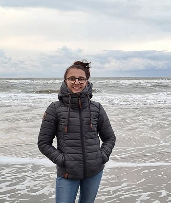 Eine junge Frau mit langen braunen, zusammengebundenen Haaren, mit Brille und in Winterjacke steht am Strand. Im Hintergrund Wellen. 