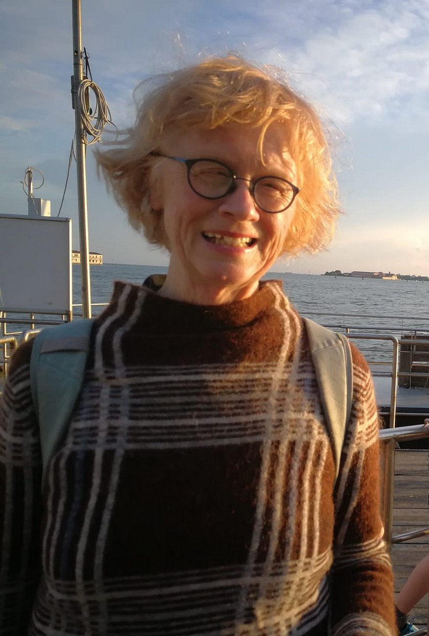 Es handelt sich um ein Portraitfoto. Zu sehen ist eine ältere Dame mit kurzen, blonden Haaren und einer Brille. Sie trägt einen braunen Wollpulli mit blau-weiß-grauen Karomuster. Im Hintergrund ist das Meer zu sehen