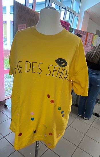 Gelbes T-Shirt mit der Aufschrift „Woche des Sehens“ und einem gemalten Auge, ausgestellt auf einer Schneiderpuppe.