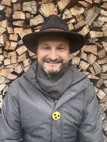 Ein Bild von Christian Ritter, ein Mann in einer grauen Jacke, einem grauen Schal und einem Filzhut. Er lächelt in die Kamera, sein Bart hat ein paar graue Stellen. An seiner Jacke trägt er ein Blindenabzeichen.