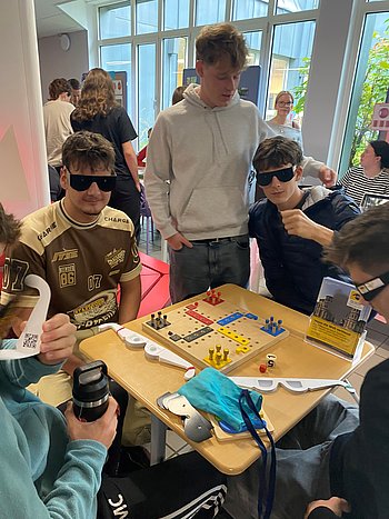 Gruppe junger Menschen spielt ein Brettspiel mit Simulationsbrillen für Sehbehinderungen.
