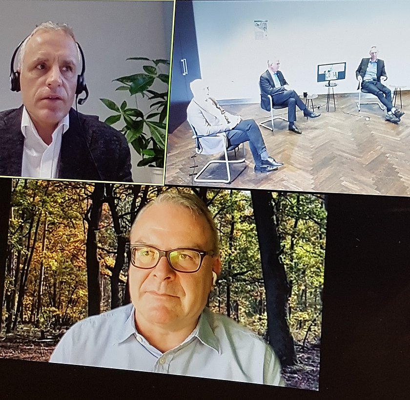 Dreigeteilter Bildschirm zeigt oben links das Porträt eines Mannes mit Headset, oben rechts drei Männer auf Stühlen sitzend, zwischen ihnen ein Tisch, auf dem ein Bildschirm steht, und unten das Porträt eines Mannes mit Brille im hellblauen Hemd, im Hintergrund ein Laubwald.