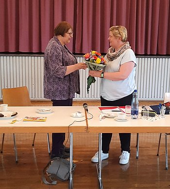 Maria Knoke (links) vom Leitungsteam überreicht Blumen an Claudia Middendorf. Sie stehen hinter zwei Tischen, auf denen eine Flasche Wasser steht und Papiere liegen. Beide haben kurzes Haar, Maria Knoke dunkles, Claudia Middendorf blondes. Im Hintergrund ist eine Bühne mit geschlossenem Vorhang zu sehen. (Foto: privat)Papi