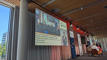 Das Podium des Raum 1 der Inklusionstage ist von der Seite zu sehen. Im Vordergrund ein Screen, auf den das Podium projiziert wird, zusammen mit Audiotranskription und Gebärdensprachdolmetschung. Die Frage "Was muss passieren, damit alle digital teilhaben können" steht im Raum (und auf dem Screen) und wird von den Gästen diskutiert.