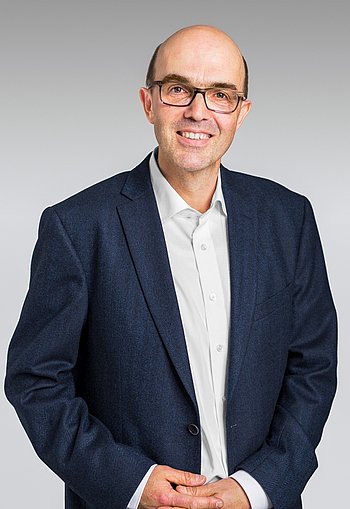 Ein Mann mittleren Alters lächelt freundlich ind die Kamera. Er trägt eine Brille, ein weißes Hemd und ein blaues Sacko.. 