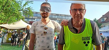 Ein junger Mann mit Simulationsbrille hält die Hand auf die Schulter eines Guides und lässt sich führen