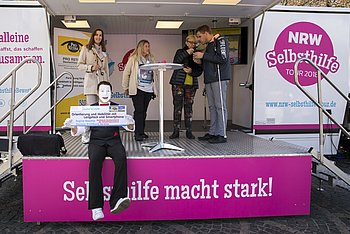 Mehrere Leute stehen mit Mikrofonen auf der Bühne eines Trucks. Unter der Bühne hängt ein Banner mit der Aufschrift "Selbsthilfe macht stark!"