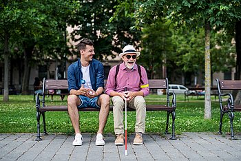 Ein junger Mann und ein älterer Mann mit Sonnenbrille, Hut und Blindenlangstock sitzen auf einer Parkbank und lachen.