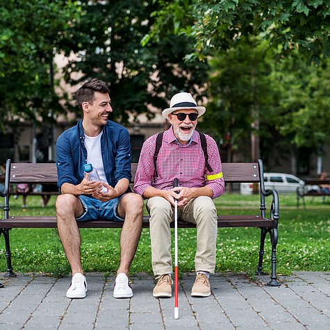 Ein junger Mann und ein älterer Mann mit Sonnenbrille, Hut und Blindenlangstock sitzen auf einer Parkbank und lachen.