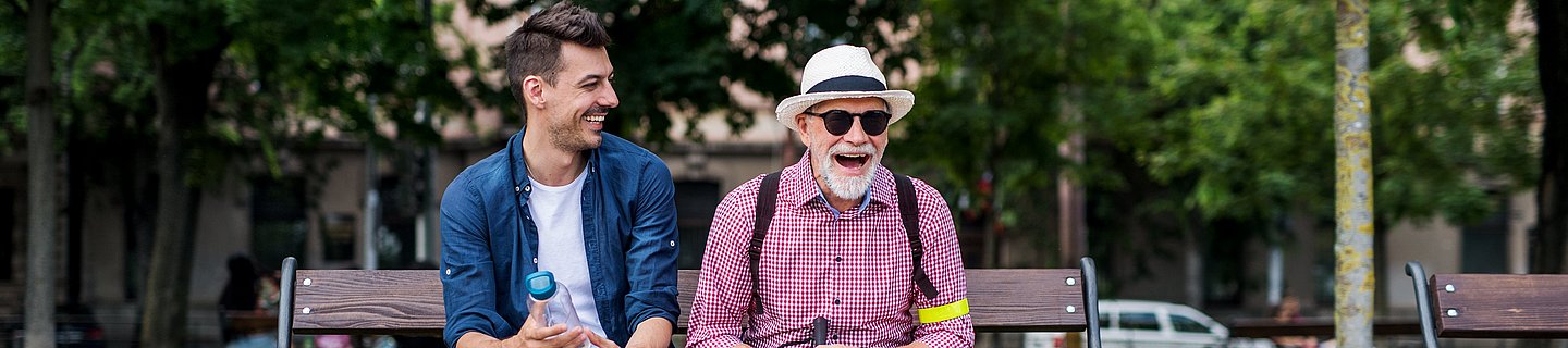 Ein junger Mann und ein älterer Mann mit Sonnenbrille, Hut und Blindenlangstock sitzen auf einer Parkbank und lachen.