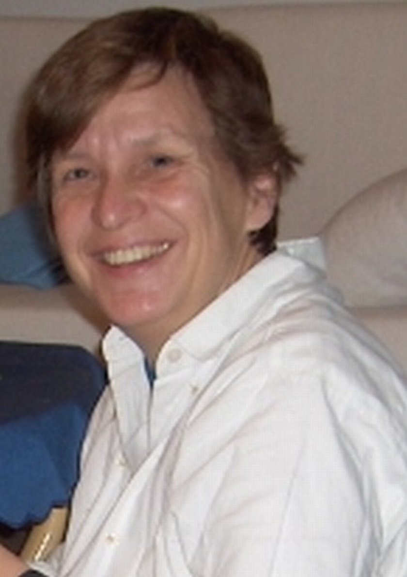 Auf dem Bild zu sehen ist eine Dame mittleren Alters. Sie hat kruze, braune Haare und braune Augen. Sie trägt eine weiße Bluse und lächelt in die Kamera. Im Hintergrund zu sehen ist eine Couch und ein Tisch mit blauer Tischdecke