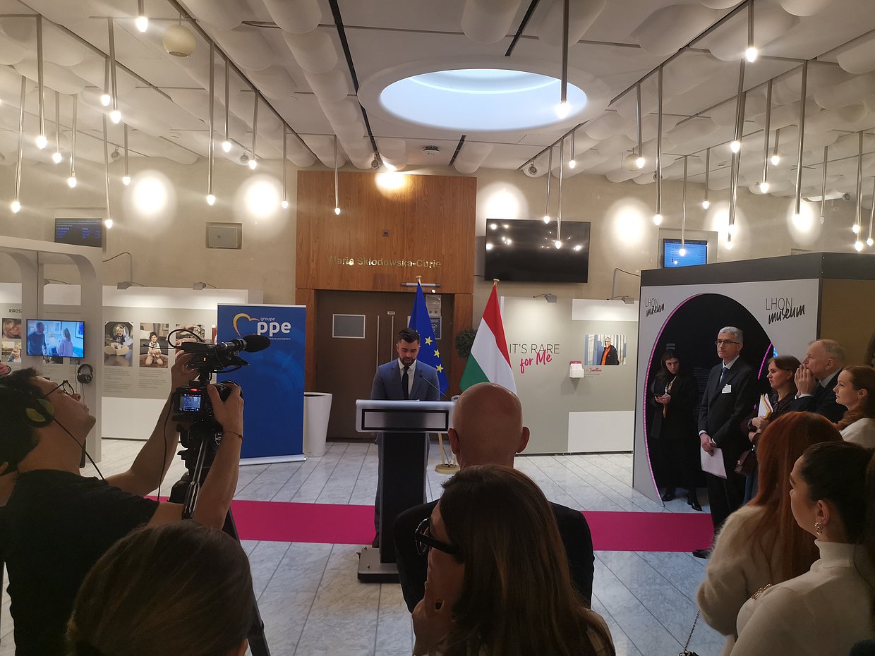 Auf dem Foto abgebildet ist folgende Szene: In einer hellen Ausstellungshalle im Europäischen Parlament in Straßburg eröffnet András Kulja, ungarischer Europaabgeordneter, die Ausstellung „It’s Rare for Me“ zum Rare Disease Day 2026. Der Abgeordnete steht am Rednerpult und spricht; hinter ihm sind die EU-Flagge und die ungarische Flagge zu sehen. Links filmt ein Kameramann die Szene, im Vordergrund sind die Köpfe von zuhörenden Gästen erkennbar. Rechts steht eine Gruppe von Gästen und verfolgt die Ansprache. An den Wänden im Hintergrund hängen Ausstellungstafeln und Fotos zu Menschen mit seltenen Erkrankungen. Ein pinkfarbener Teppichstreifen verläuft quer durch den Vordergrund hinter dem Rednerpult entlang und führt zum Eingang des „LHON Museums“ – gestaltet als schwarzer Tunnel, der in regelmäßigen Abständen von pink leuchtenden Linien akzentuiert wird.