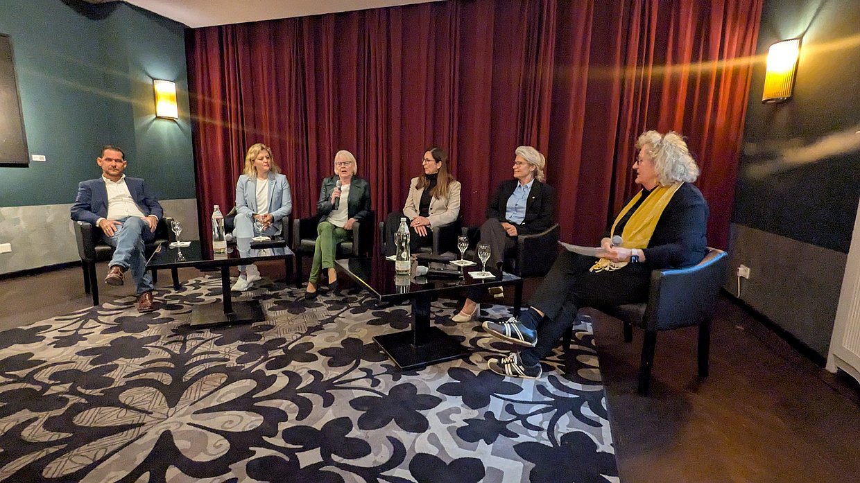 6 Personen sitzen in einem Halbkreis in einer gemütlichen Wohnzimmeratmosphäre