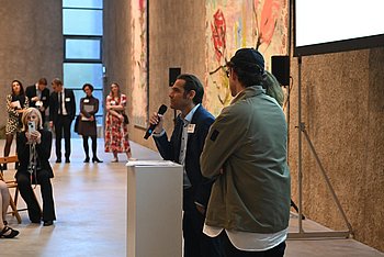 Am Rednerpult steht Dario Madani, Geschäftsführer von PRO RETINA, bei der Kunstauktion in der König Galerie in Berlin, neben ihm, mit dem Rücken zum Betrachter, der Galerist Johann König. Im Hintergrund sieht man an einer hohen Wand ein buntes Gemälde und einige Besucherinnen und Besucher der Versteigerung.