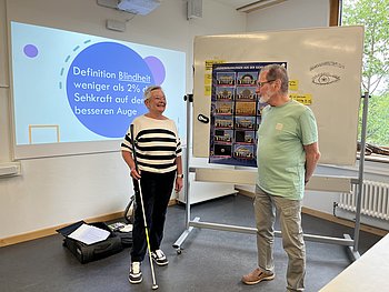 Zwei Referierende stehen in einem Klassenraum vor einer Präsentation zum Thema Blindheit und Sehbeeinträchtigungen. Eine Frau hält einen Langstock, daneben ist ein Poster zu Augenerkrankungen zu sehen.