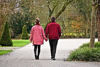 Ein Mann und eine Frau gehen Hand in Hand einen ruhigen Weg in einem Park entlang. Beide tragen Jacken, und im Hintergrund sind Bäume und Grünflächen zu sehen.