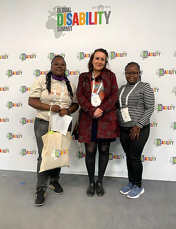 Von links nach rechts stehen: Ruby Loretta aus Uganda von der Organisation “Diverse Empowerment Foundation”, Zehra Wellmann-Sam politische Referentin der PRO RETINA, Phylis Mbeke Ndolo aus Kenia von der Organisation “Woman in Global Health” vor der Fotowand des Global Disability Summit. Das bunte Logo des Summits zusammen mit den Umrissen der Kontinente ist im Hintergrund zu sehen.
