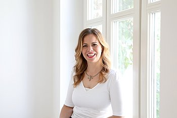 Portrait einer lächelnden Frau mit langem, blondem, gewellten Haar, braunen Augen, rotem Lippenstift und einem weißen Shirt.