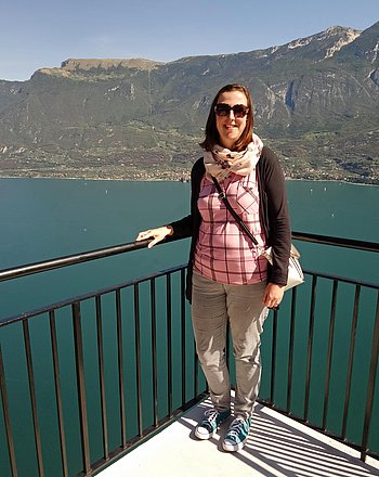 Linda Meschke Eine junge Frau schulterlangem braunem Haar und Sonnenbrille lehnt an ein Balkongitter. Im Hintergund Meer und Berge.