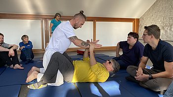 Eine Person in weißer Kleidung führt eine Selbstverteidigungstechnik mit einer Person im gelben Shirt vor, die auf dem Rücken liegt. Die anderen Teilnehmenden sitzen aufmerksam daneben und beobachten die Übung.