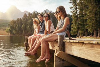 Junge Menschen sitzen im Sommer auf einem Steg an einem See. Im Hintergrund sind Bäume und Berge zu sehen.