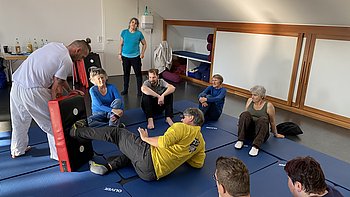 Das Bild zeigt eine Gruppe von Menschen in einem Trainingsraum mit blauen Matten auf dem Boden. Einige Personen sitzen oder liegen auf den Matten, während ein Mann vorne mit einem Schlagpolster zu sehen ist. Die Atmosphäre wirkt konzentriert, aber locker, und die Teilnehmenden hören aufmerksam zu oder beobachten die Übung.