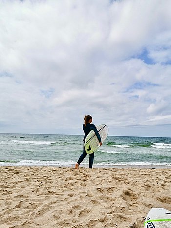 Eine Frau in Neoprenanzug steht mit einem Surfbrett in der Hand am Strand und schaut auf das wellige Meer. Sie ist von hinten zu sehen. 