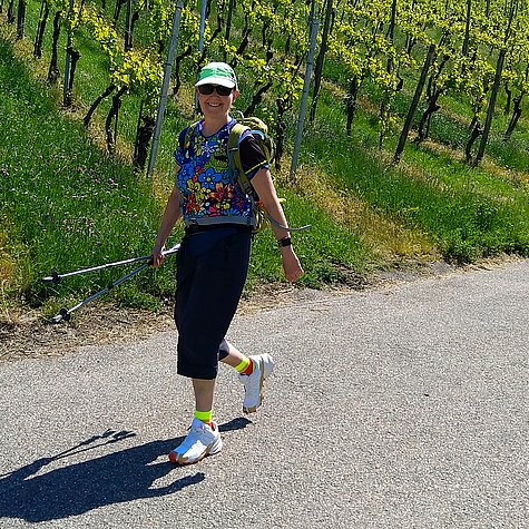 Junge Frau in Sportbekleidung mit Kappi, Sonnenbrille, Rucksack und Wanderstöcken läuft auf einer Straße an einem Weinberg entlang leicht bergab.
