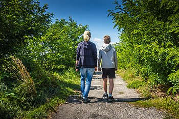 mother and son hiking Eine Frau mit blondem Pferdeschwanz und ein Junge mit kurzem braunen Haar gehen einen Weg entlang, der rechts und links von grünen Bäumen und Büschen flankiert ist. Man sieht sie von hinten.