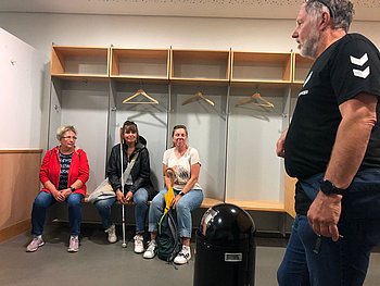 Drei Frauen sitzen in der Umkleidekabine der Gastmannschaft. Über ihren Köpfen sind Kleiderbügel zu sehen, die an der Wand hängen. Am rechten Bildrand steht Stadionführer Bernhard