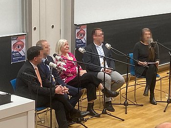 Zwei Frauen und drei Männer sitzen in einer Reihe auf einer Bühne, jeder hat einen Mikrofonständer vor sich.
