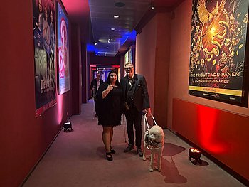 Von links nach rechts: Dr. Sandra Jansen (Fachreferentin für Diagnose & Forschung) und Jörg-Michael Sachse-Schüler (Finanzvorstand von PRO RETINA) stehen gemeinsam in einem langen Flur des historischen Colosseum Filmtheaters in Berlin. Jörg-Michael Sachse-Schüler wird von seinem Blindenführhund / Assistenzhund begleitet, der vor ihm steht. Der Flur ist stimmungsvoll beleuchtet. Die Wände sind in roten Farbtönen gehalten. An den Wänden sind Filmplakate angebracht. Diese Plakate werden von zwei Leuchten beleuchtet, die auf dem Boden stehen. 