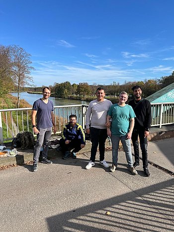 Alternativtext: Fünf junge Männer stehen und hocken nebeneinander auf einer Brücke bei sonnigem Wetter. Im Hintergrund ist ein ruhiger Fluss, umgeben von Bäumen mit Herbstlaub, und ein klarer, blauer Himmel zu sehen. Alle lächeln in die Kamera, die Stimmung ist entspannt und fröhlich.