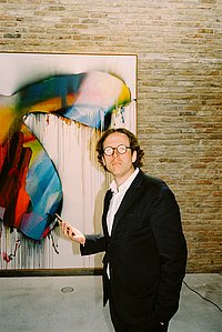 Johann König steht in seiner Galerie vor einem abstrakten Gemälde in blau-weiß-roten Farben, das an einer rohen Klinkerwand hängt. Er trägt einen dunklen Anzug und Brille und blickt im Halbprofil in die Kamera.