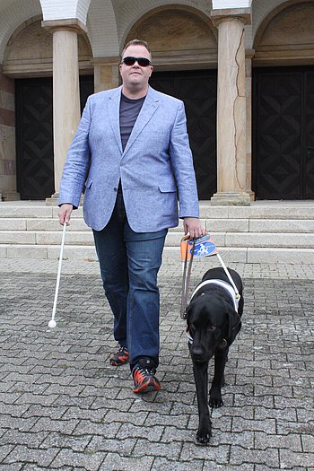 Wolfgang Schweinfurth wird von seinem schwarzen Blindenführhund geführt. Der Labrador trägt ein weißes Führgeschirr, das Wolfgang Schweinfurth in der linken Hand hält.