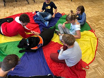 Kinder sitzen mit einem Blindenführhund auf einem bunten Tuch 